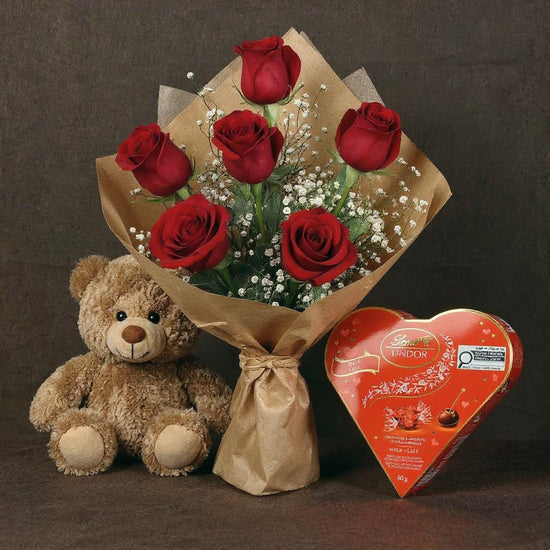 Bouquet of red roses, teddy bear, and Lindt heart-shaped chocolate on a dark background