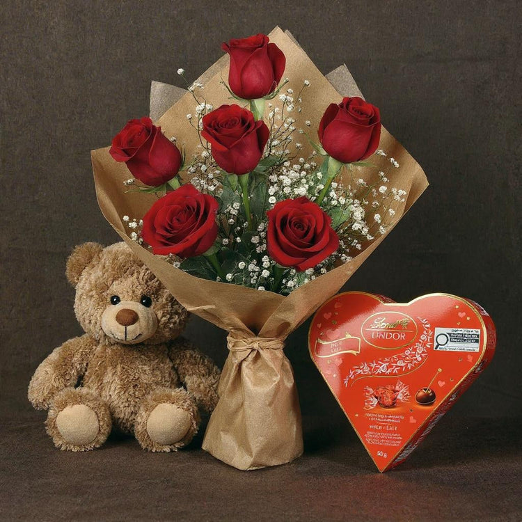 Bouquet of red roses, teddy bear, and Lindt heart-shaped chocolate on a dark background